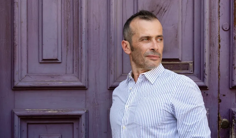 Foto ritratto di Andrea in piedi davanti a una porta. Ad Andrea è stata diagnosticata la sclerosi multipla recidivante remittente e ha avuto problemi sia alla vescica che all'intestino. 